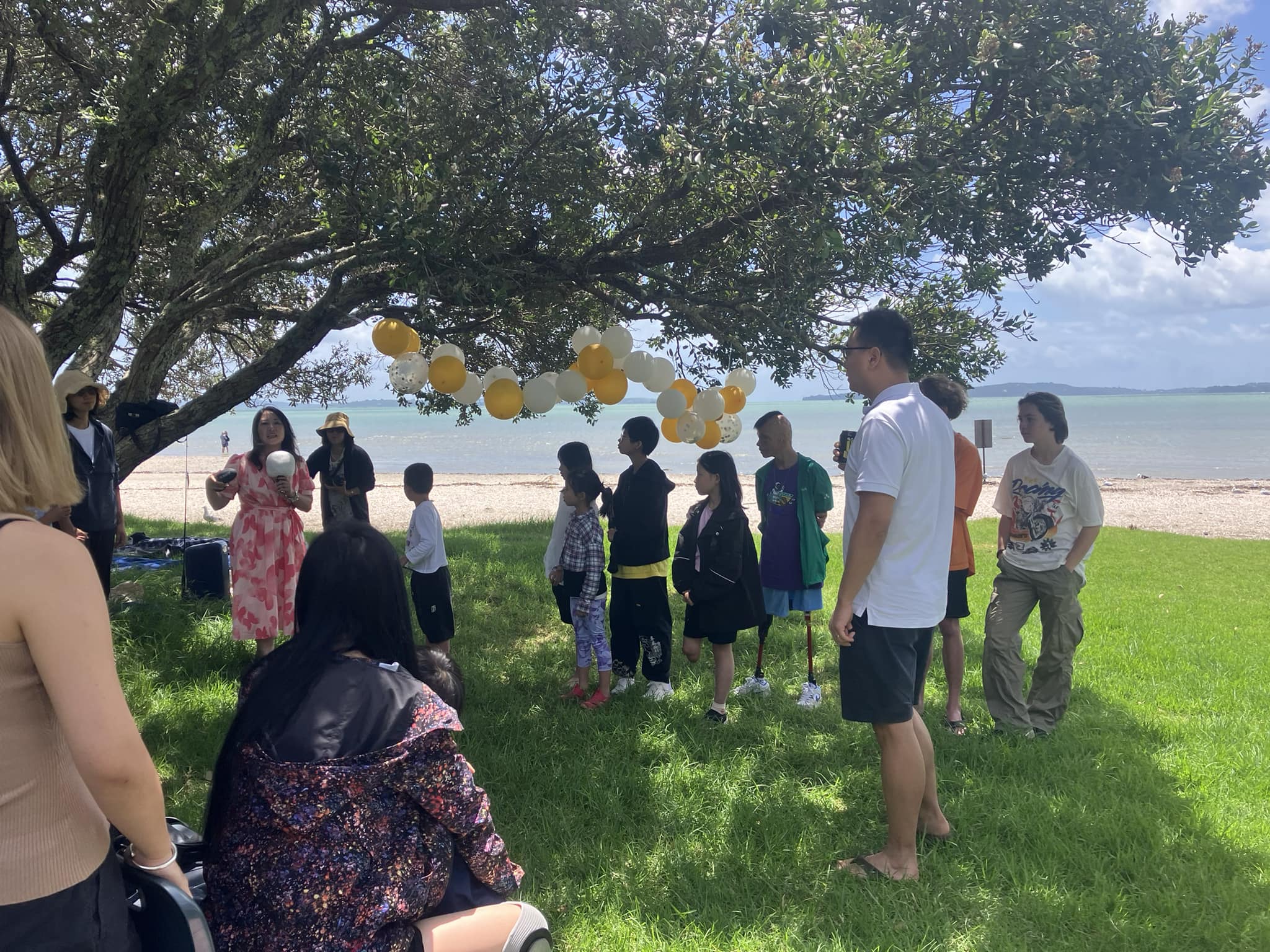 Auckland Anniversary Day celebration: People gather under a tree near the beach with balloons.