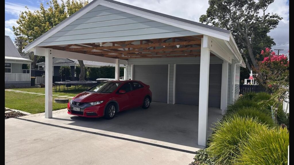 Exterior shot of a modern carport next to a garage, with a red car parked inside. Features light blue siding and white trim.